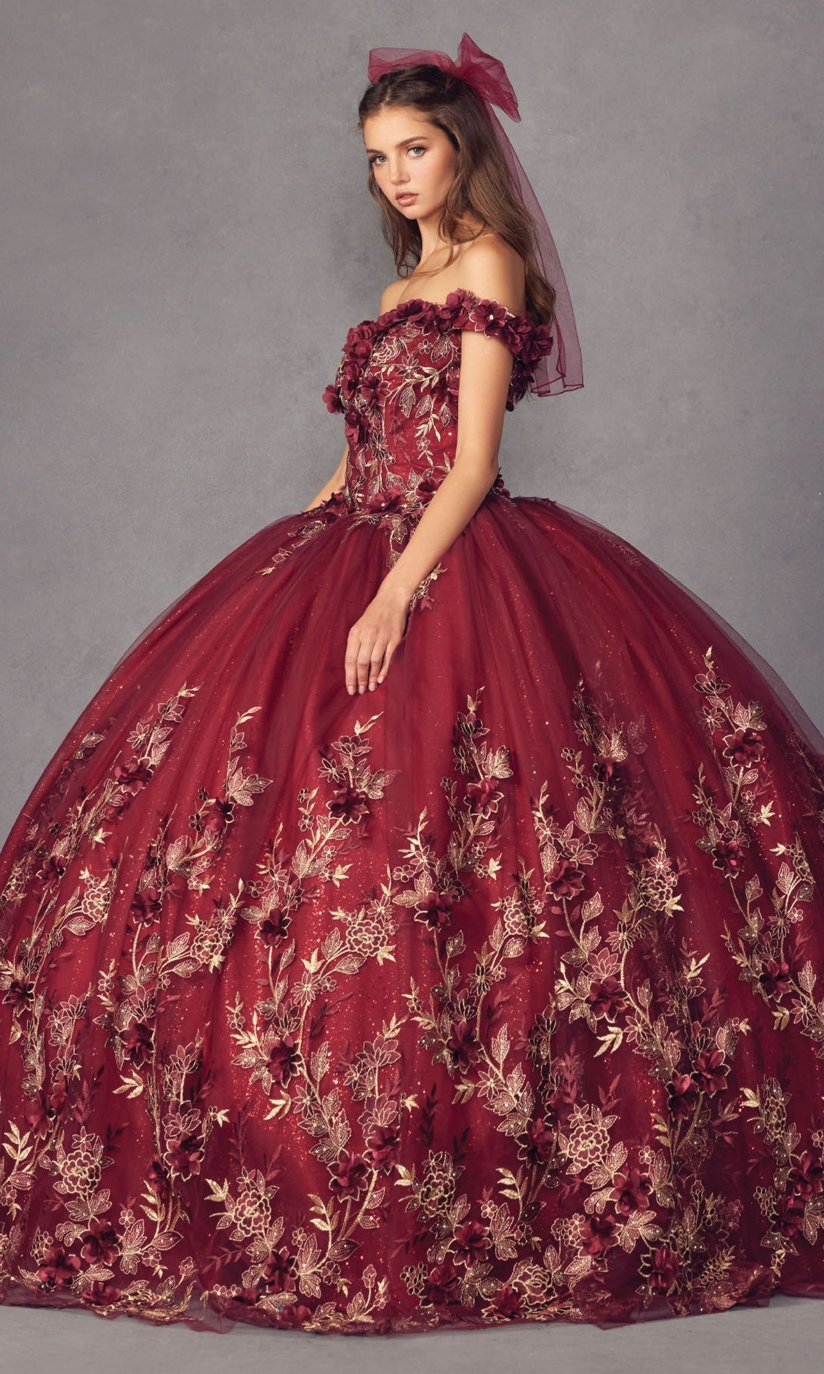 A woman poses in the Juliet 1447 Off-the-Shoulder Quinceañera Dress, a burgundy glitter ball gown with gold floral embroidery and a matching hair bow, set against a plain gray background.